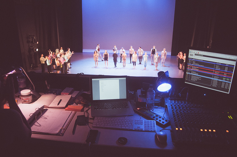 Photo: Harry Butcher. A typical QLab setup for stage sits high in the stands as a group of performers rehearse on stage