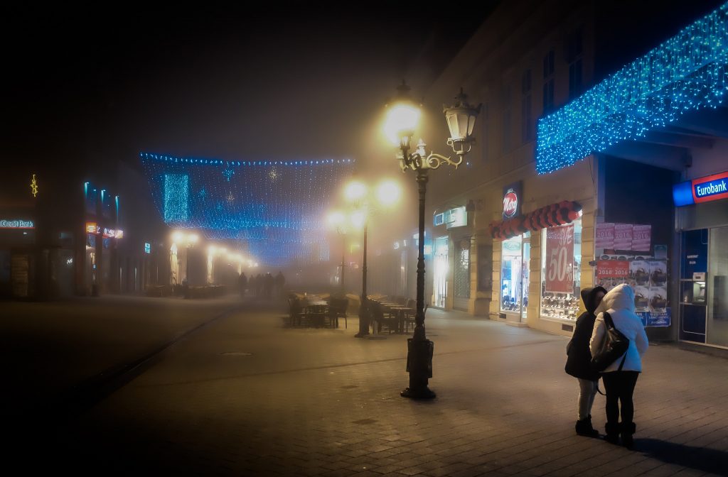 Two women kiss on a foggy European destination street with blue and white light displays, secluded from the others walking down the road. Article written by Adriane Kuzminski.