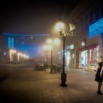 Two women kiss on a foggy European destination street with blue and white light displays, secluded from the others walking down the road.