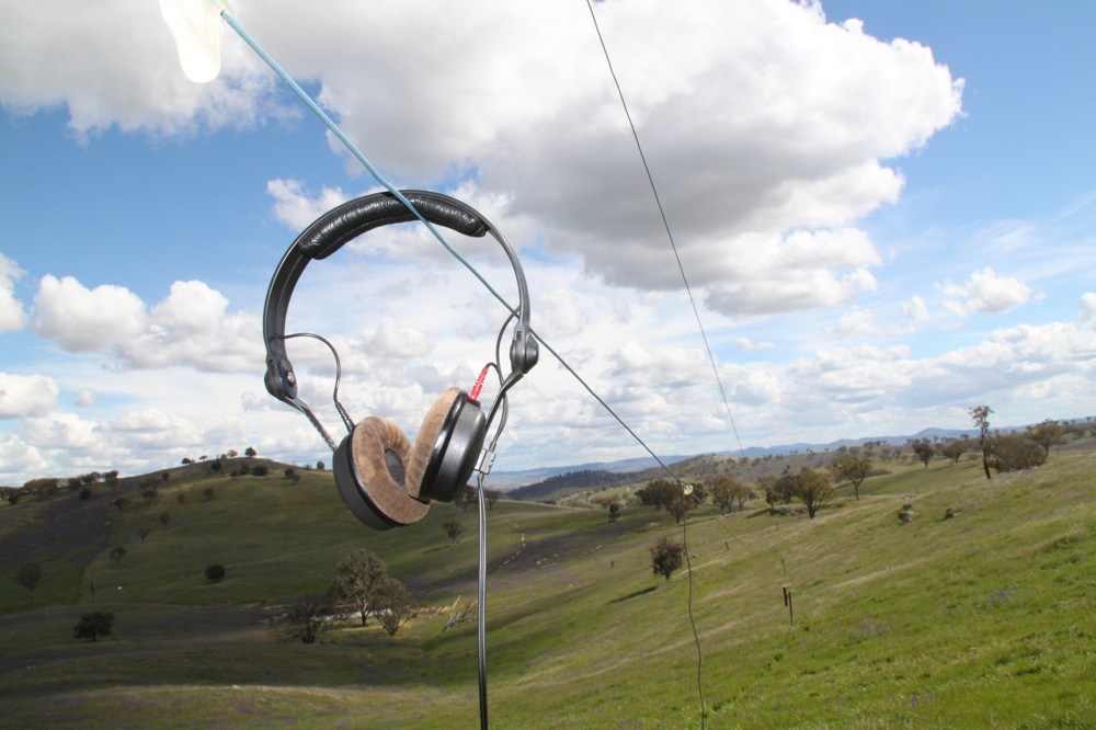 A pair of headphones hang on a long line in a rolling field. Article written by Adriane Kuzminski.