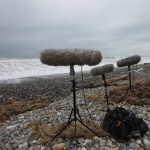 Three boom mics sit on a rocky beach facing a choppy sea underneath a blanket of dark clouds.