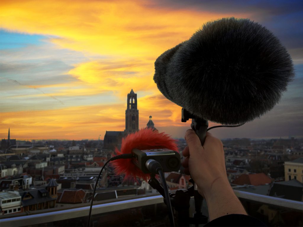 A boom mic and digital recorder sit at the edge of a rooftop facing a dusk sky over a city in the Netherlands. Article edited by Adriane Kuzminski.
