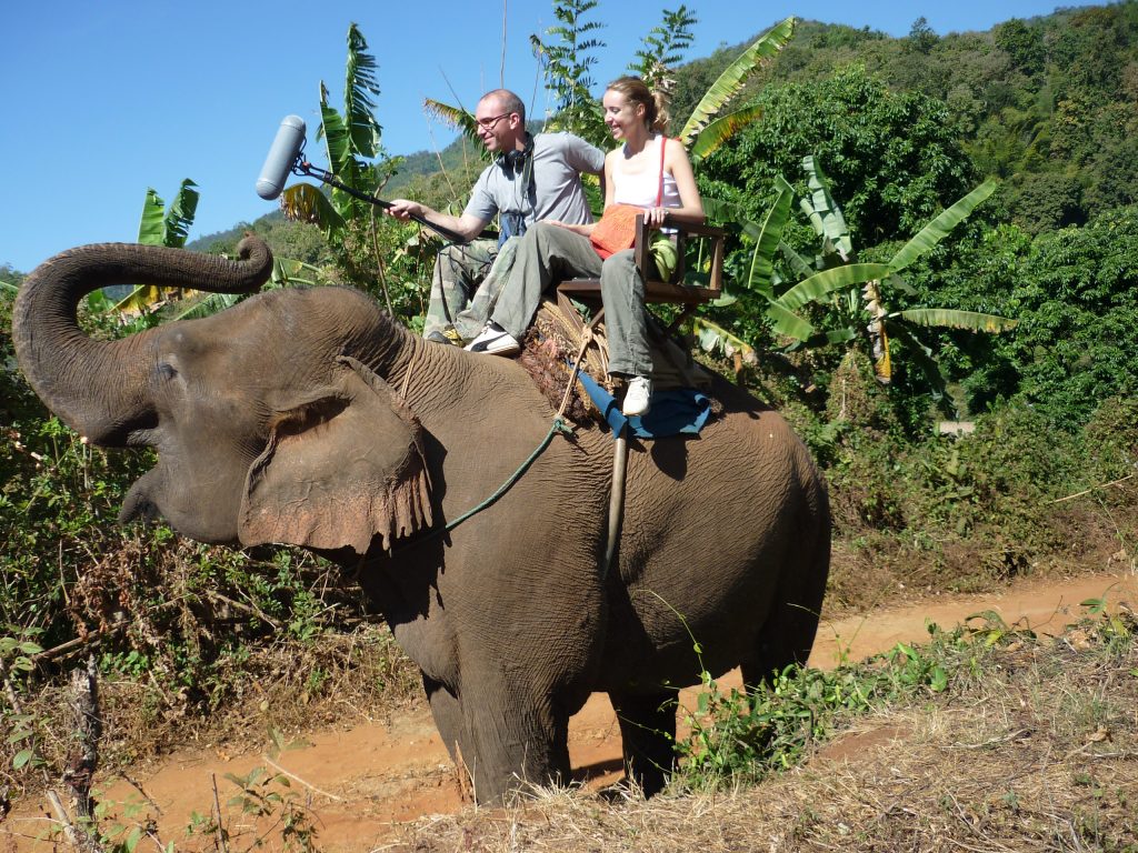 Two people ride an elephant in Thailand. The man holds a boom mic near the elephant's head as it trumpets. Article edited by Adriane Kuzminski.