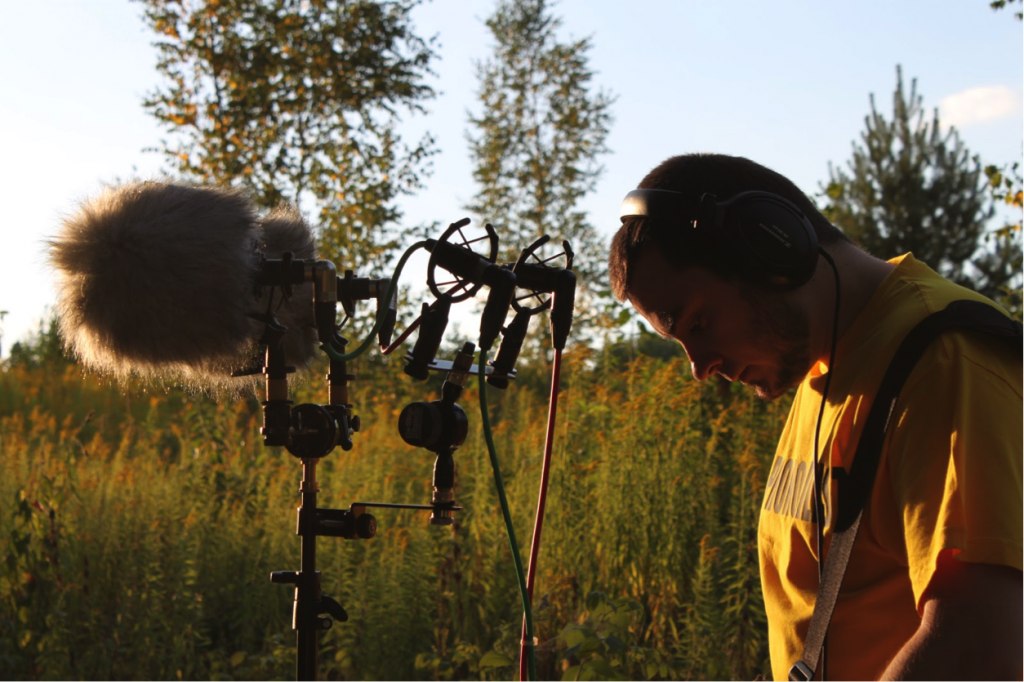 Serj stands next to his mic, listening with headphones, in the middle of a field. Article edited by Adriane Kuzminski.