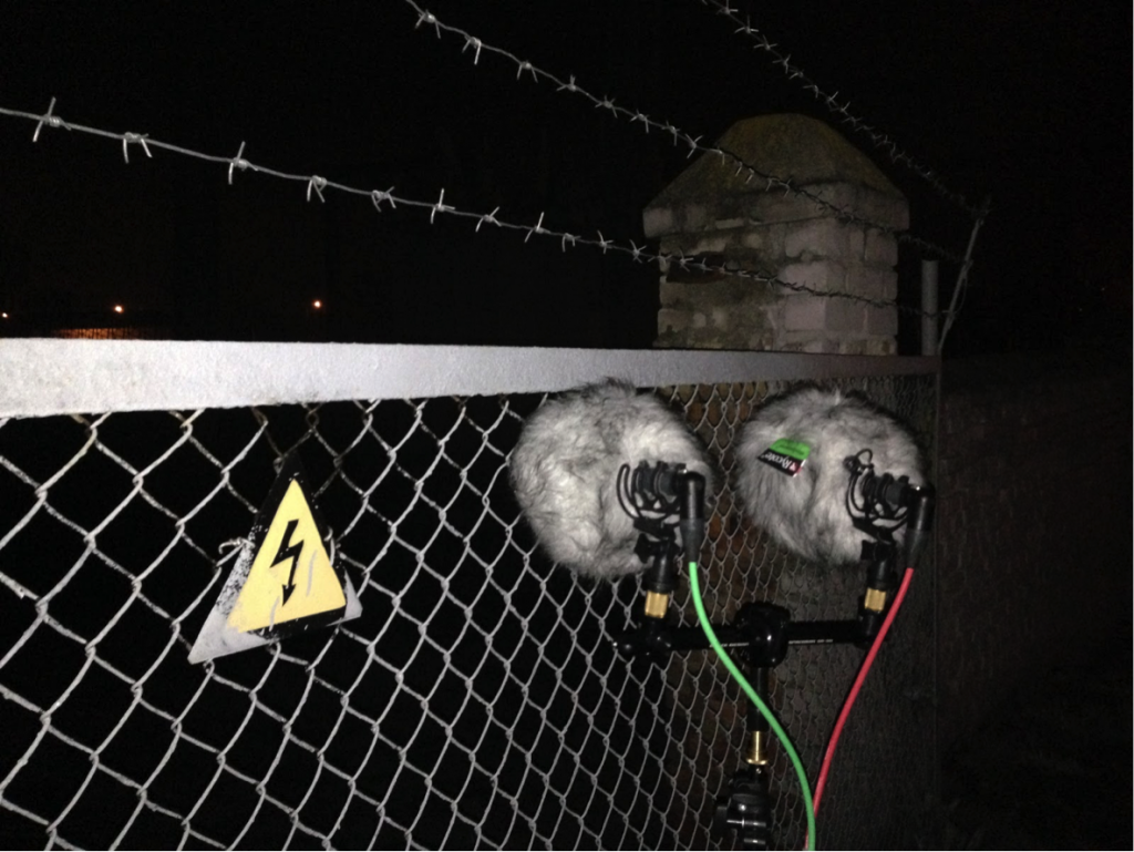 A pair of mics face a buzzing electric and barbed-wire fence.
