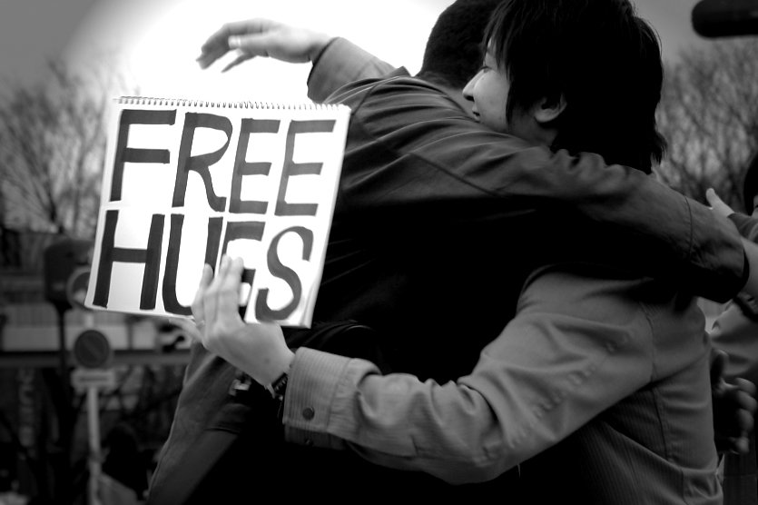 Two people hug with one person holding a free hugs sign. Article written by Adriane Kuzminski.